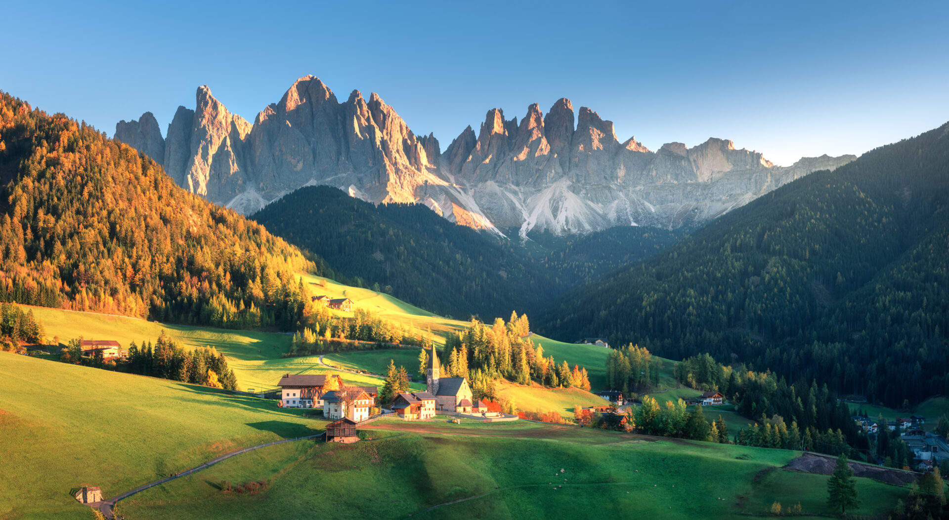 luxury hiking tours: Landscape with village with houses, church, green meadows, trees, rocks, blue sky in autumn in the Dolomites, Italy.