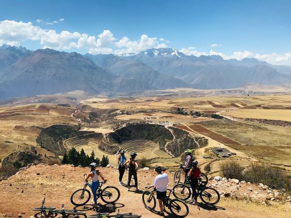 A group of six mountain bikers standing with their bikes overlook with the circular terraces of the Moray ruins in the Sacred Valley of Peru.